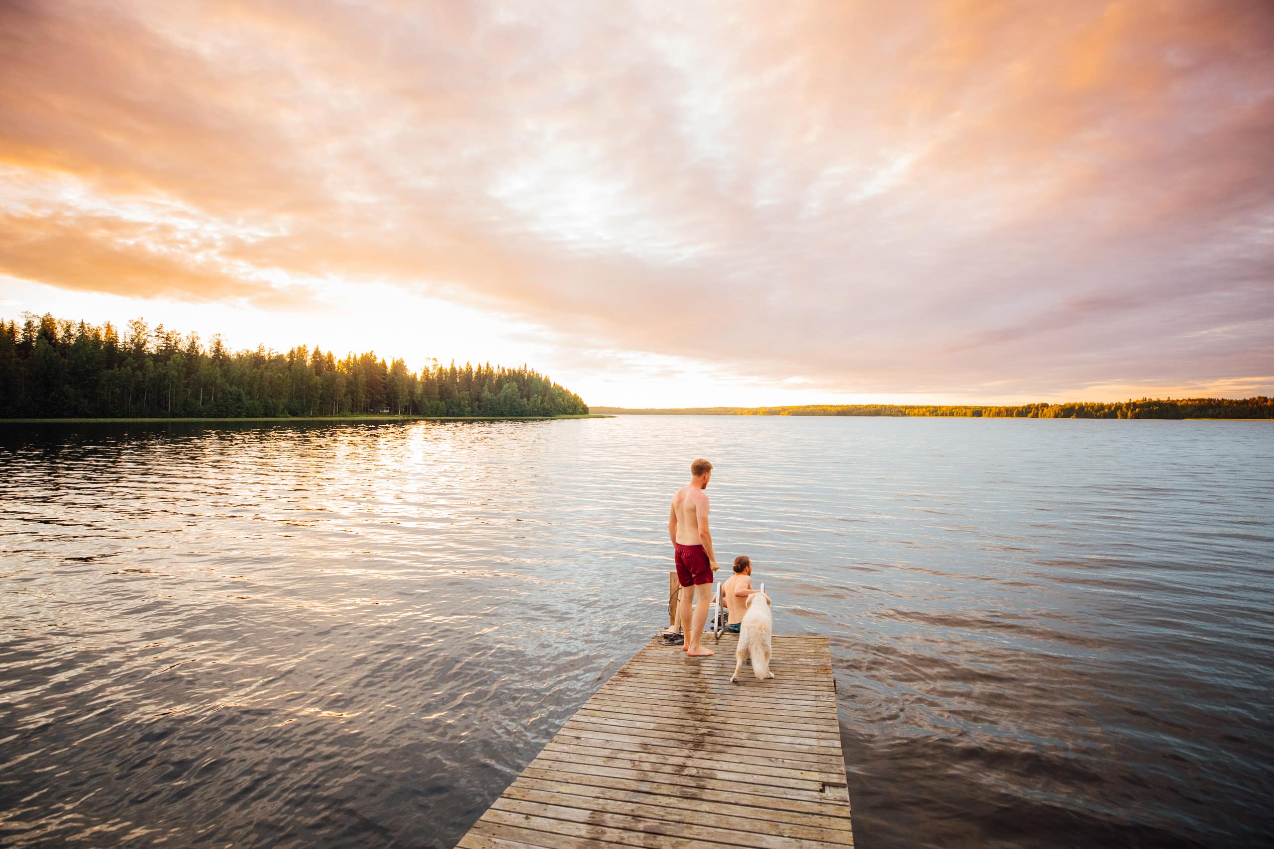 Two persons and a dog on a wooden deck over a lake.