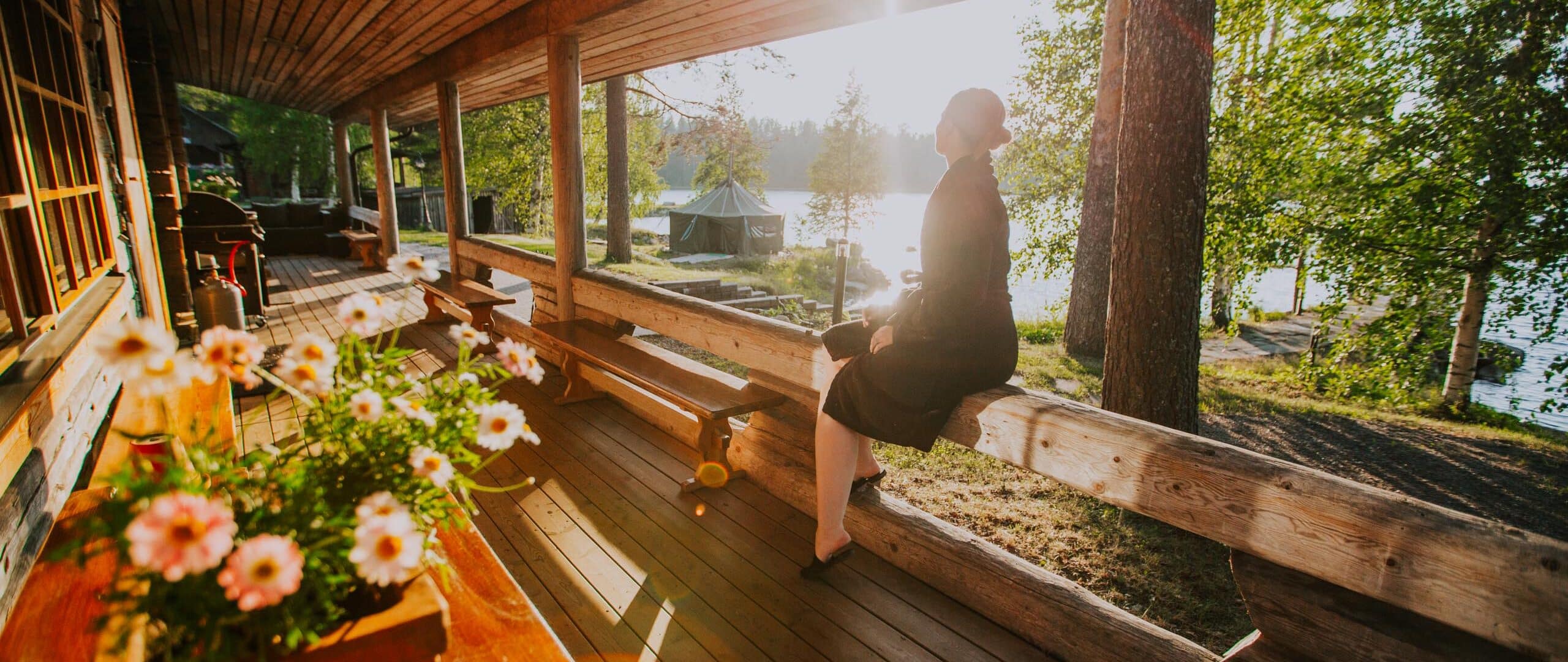 Person sitting on a railing on a terrace. There are flowers on the terrace and lake and sunshine in the background.