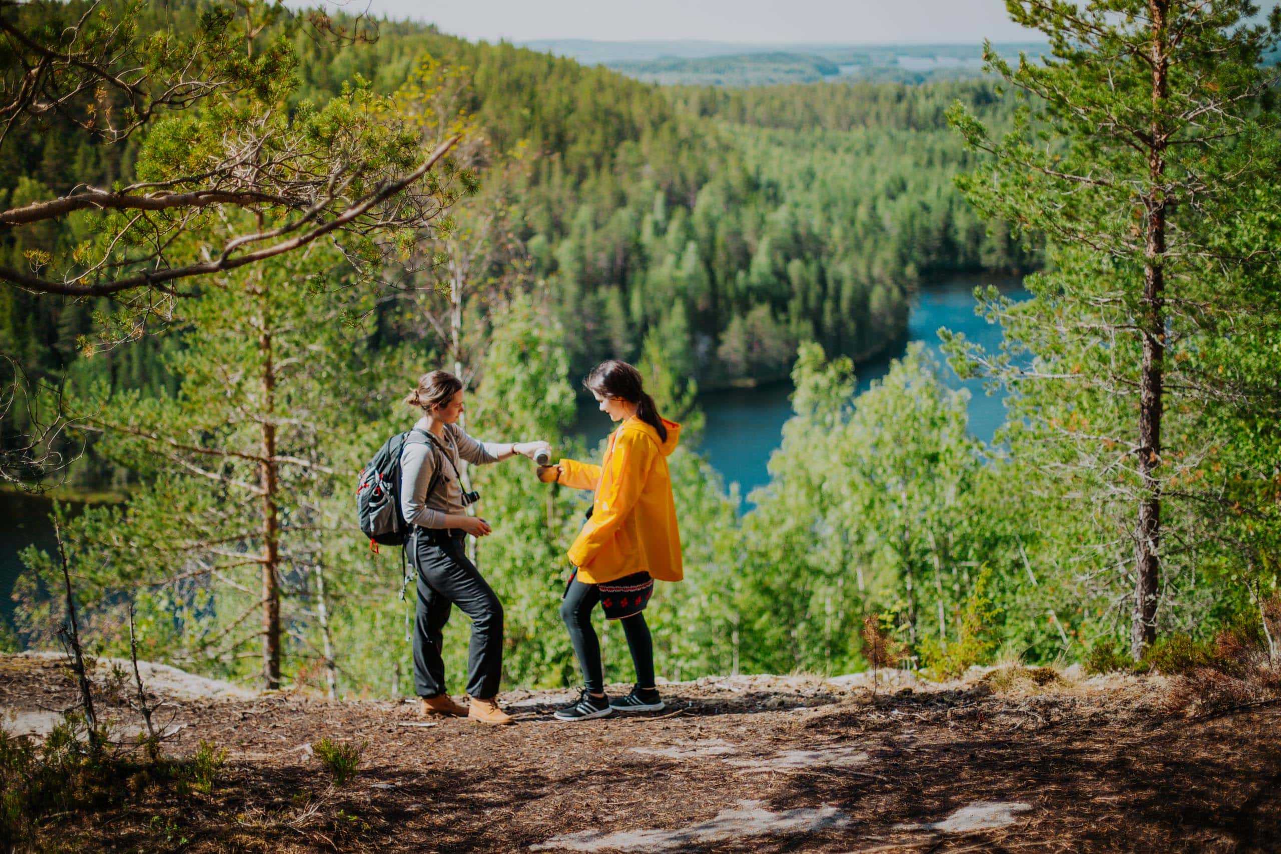 Two persons standing on a viewpoint. There is a forest and lake below.
